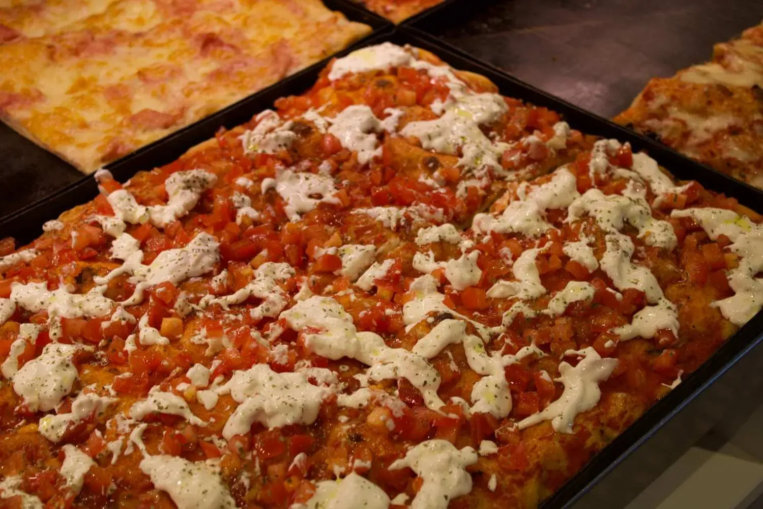 Multiple slices of authentic Roman pizza al taglio topped with fresh mozzarella, herbs, and tomato sauce displayed on black trays at a traditional Roman pizzeria.