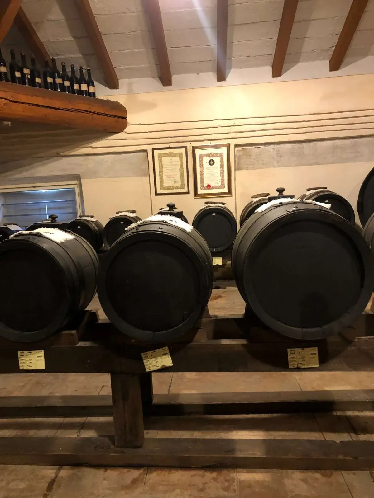 Large black wooden barrels used for aging balsamic vinegar in a traditional historic cellar with exposed wooden beams and framed certificates on the walls, representing authentic Emilia-Romagna balsamic vinegar production.
