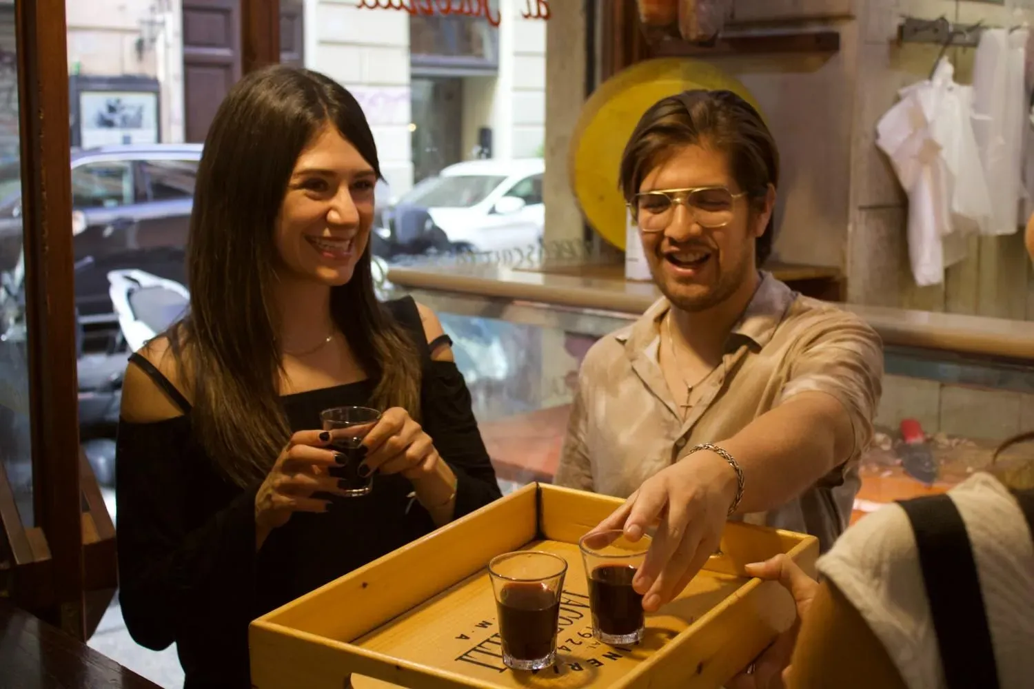 A couple toasting with glasses of limoncello during a Rome food tour, with a wooden tasting board of cheese cubes visible on the table.