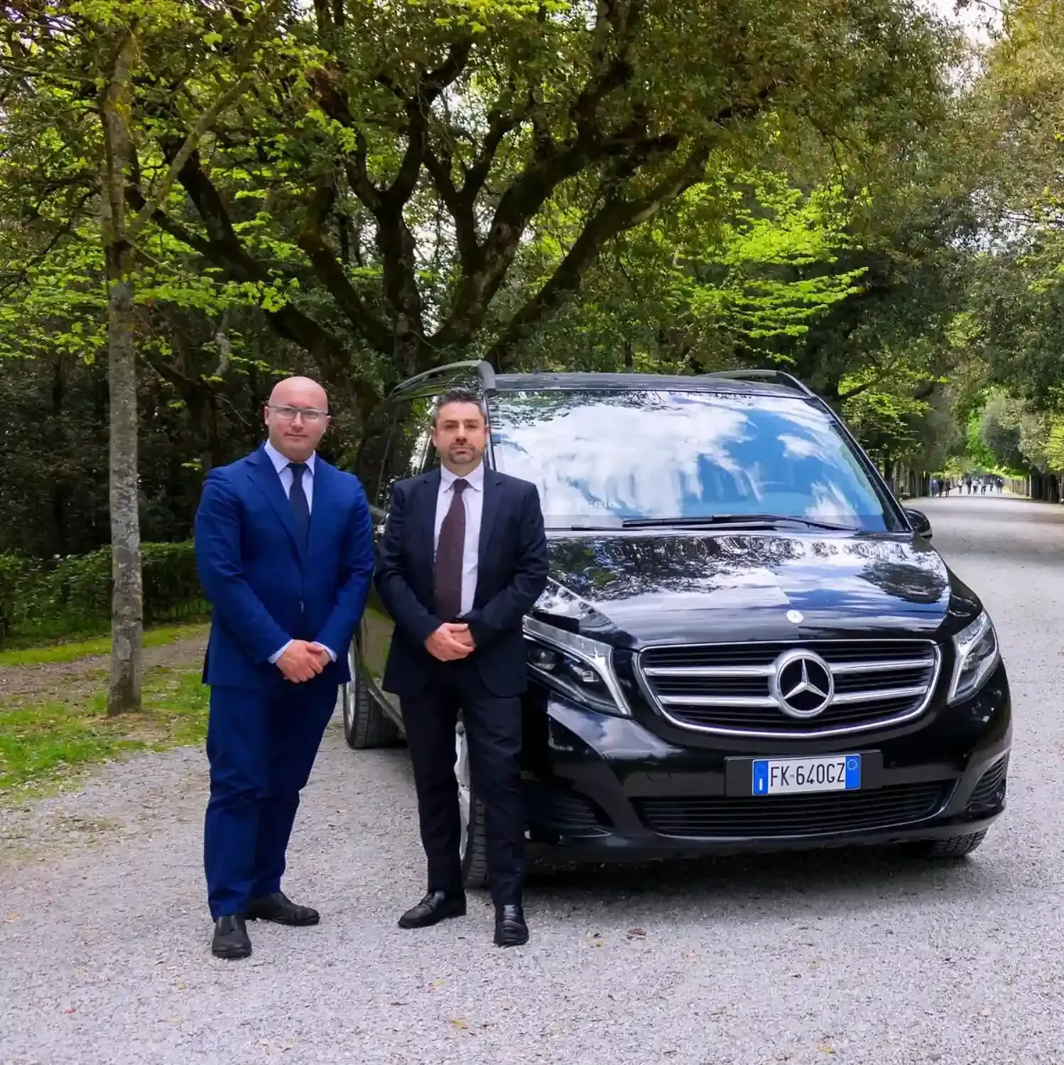 Two professional drivers standing with black Mercedes luxury SUV in tree-lined Tuscan countryside for premium tour experience