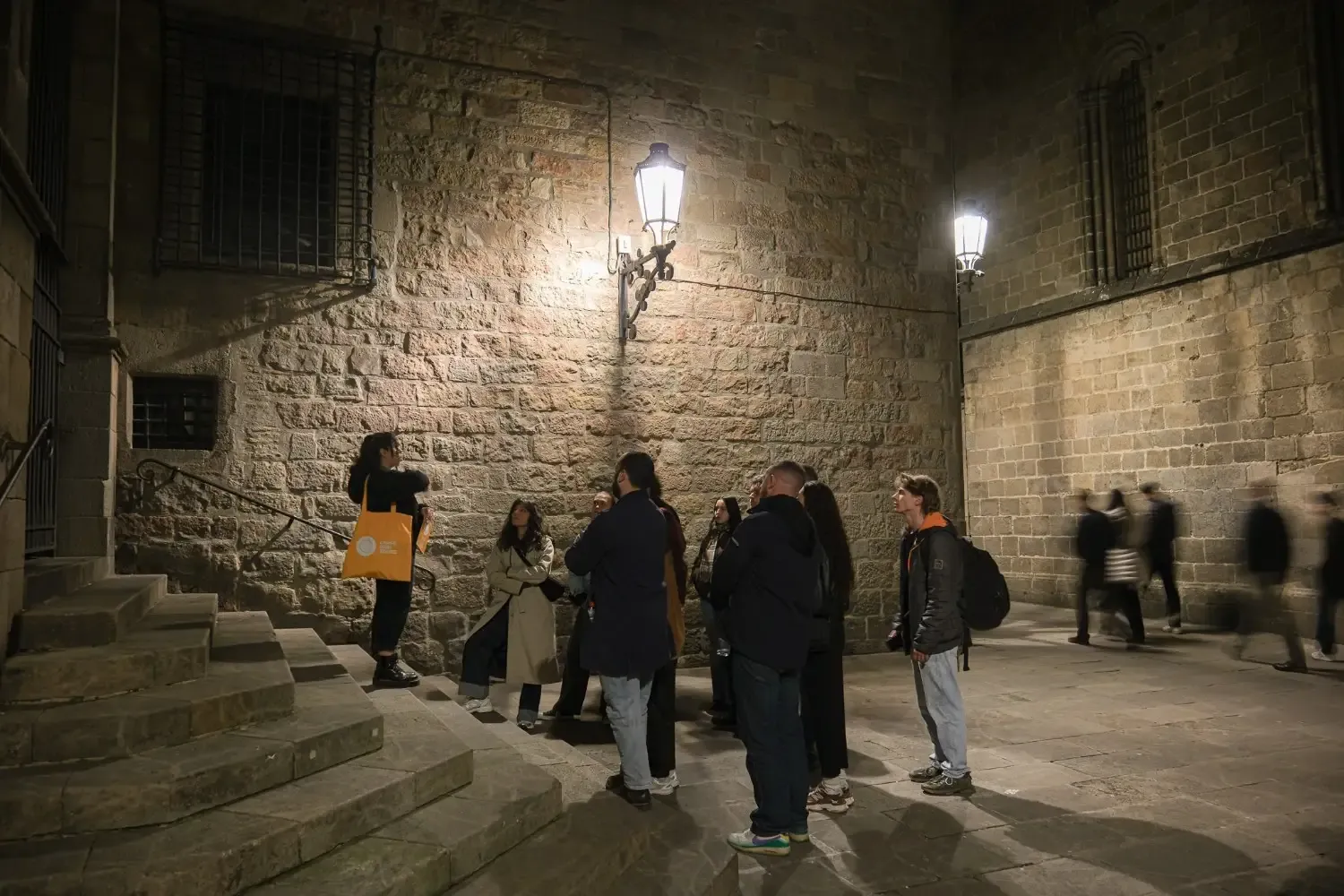 Ghost tour participants standing on historic stone steps in El Born neighborhood with dramatic stone walls and archways illuminated at night, exploring the paranormal past of Barcelona.