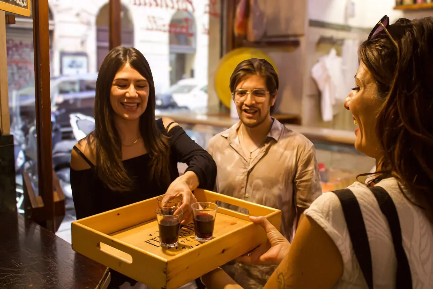 A group of smiling food tour participants enjoying limoncello shots together on a charming Rome street with historic buildings and evening lighting in the background.
