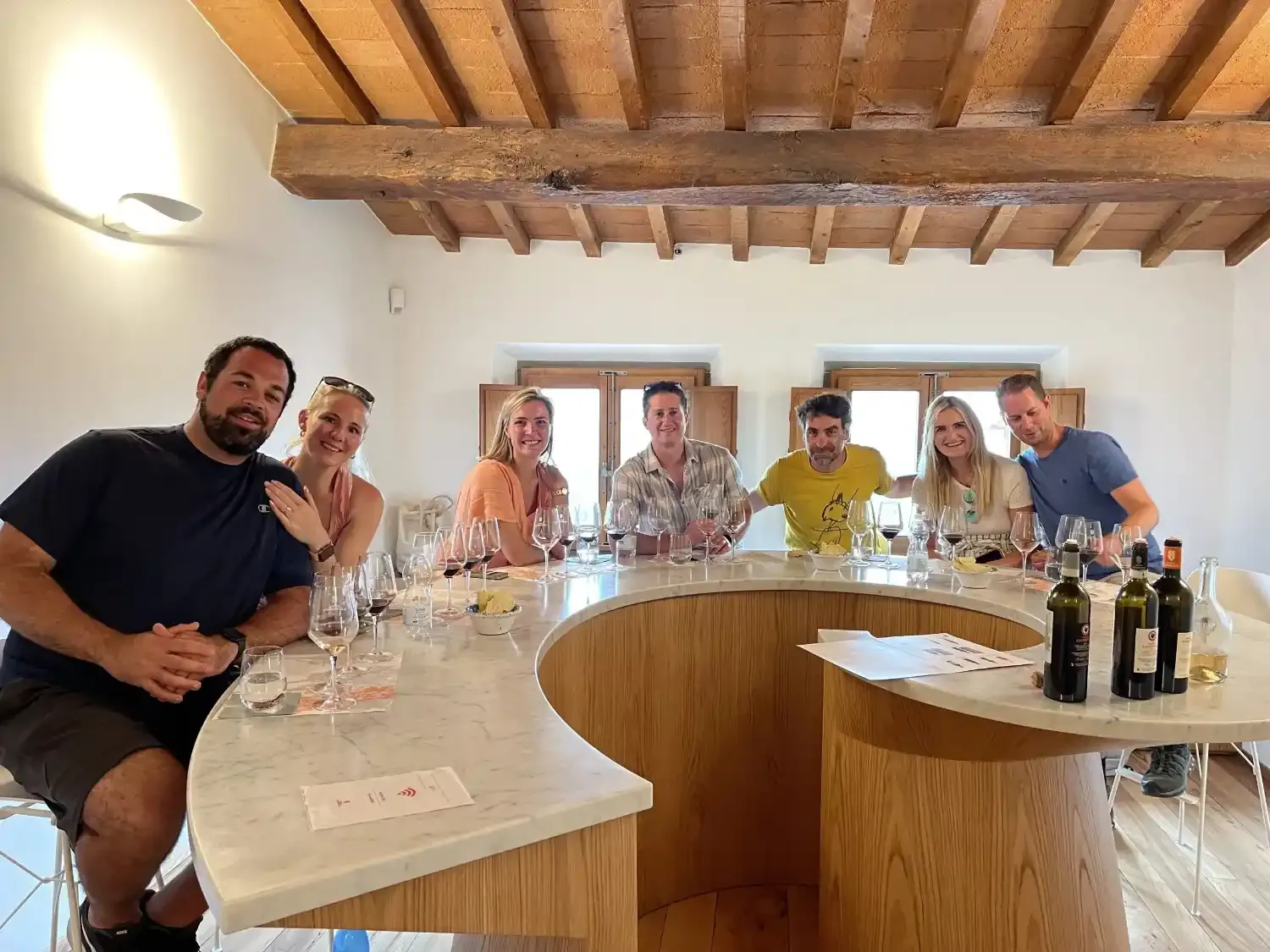 A group of happy tourists sitting around a modern, curved marble bar during a guided wine tasting at a winery in Chianti.