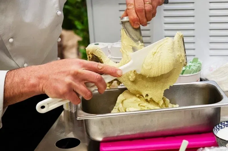 A participant's hands working with pistachio-colored gelato mixture in a metal container during a traditional gelato-making masterclass in Florence, learning from an artisan instructor.