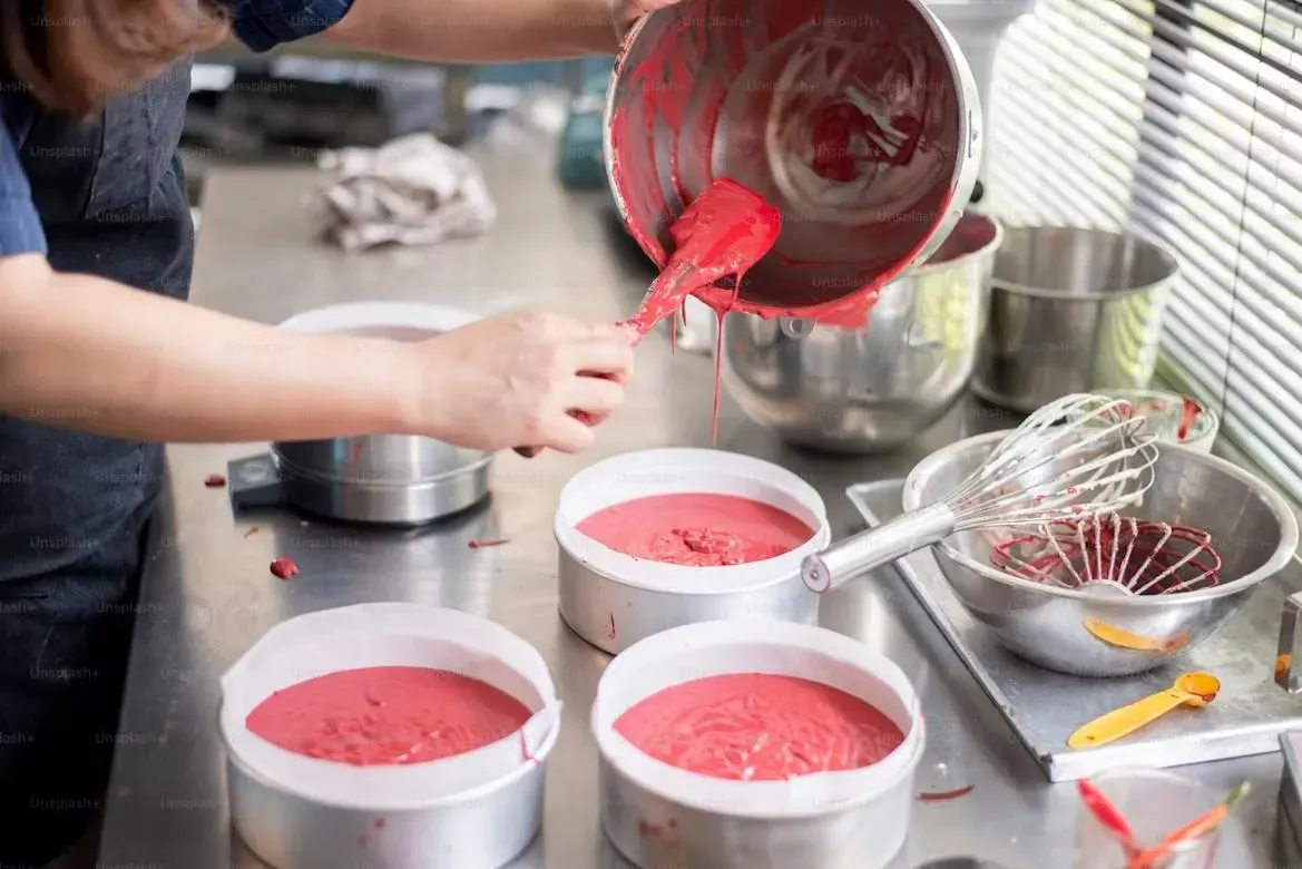 A chef pouring vibrant pink gelato or sorbet mixture from a bowl into white containers during a traditional Italian gelato-making cooking class, with whisks and measuring tools visible on the counter.
