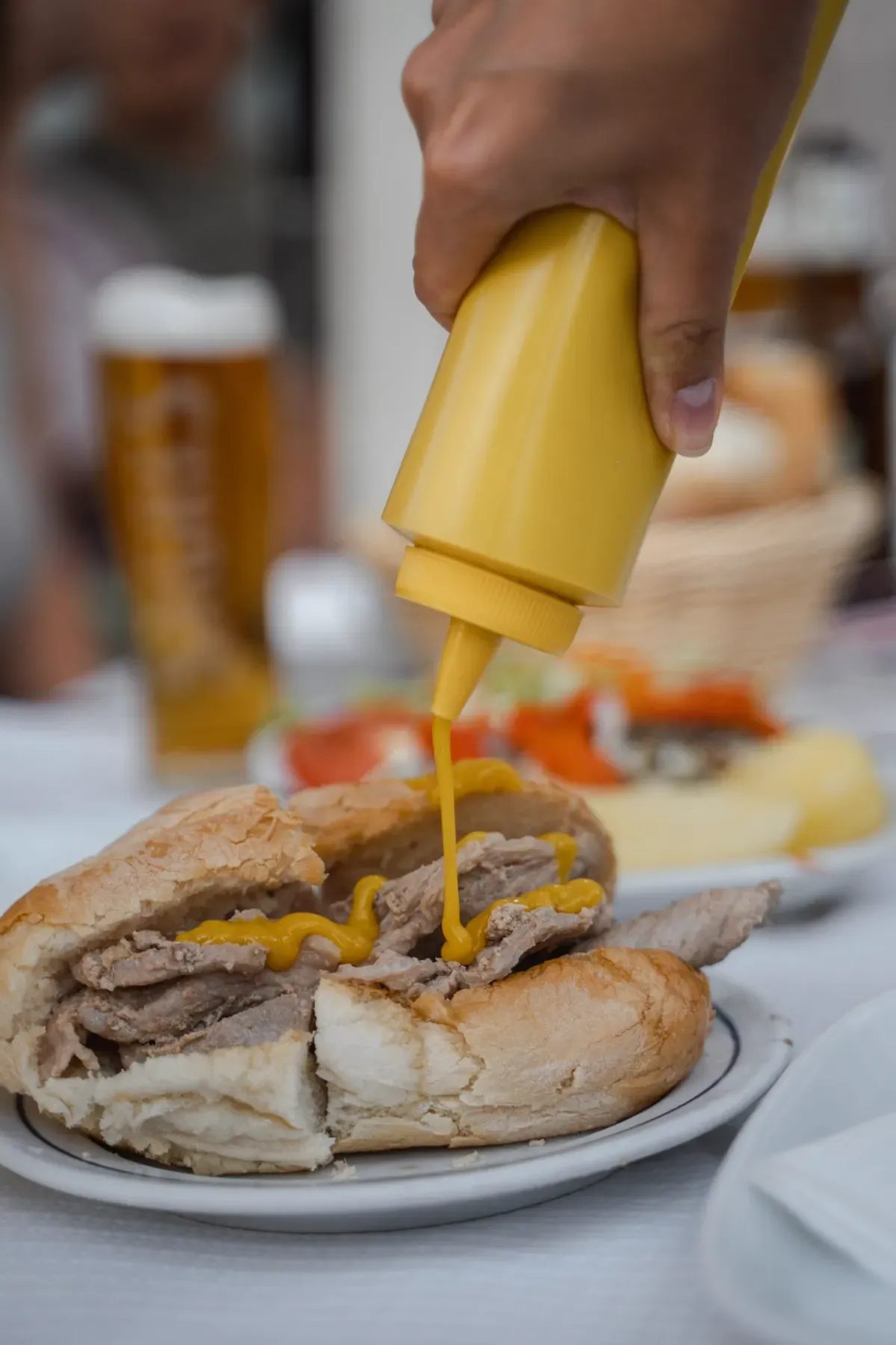 Close-up of a hand squeezing a yellow mustard bottle onto a traditional Portuguese bifana sandwich with sliced pork on a white plate, with a blurred glass of beer in the background.