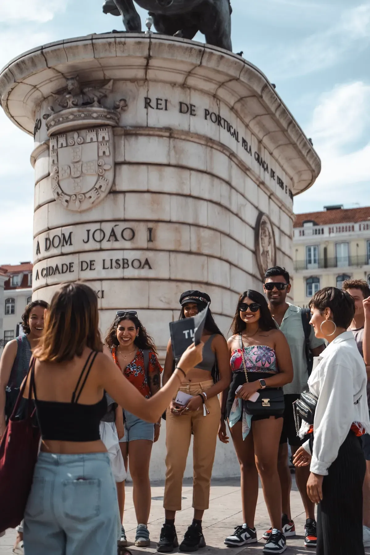 Group of young, diverse tourists on a tipsy tour in Lisbon, standing in front of the statue of Dom João I in Praça da Figueira.