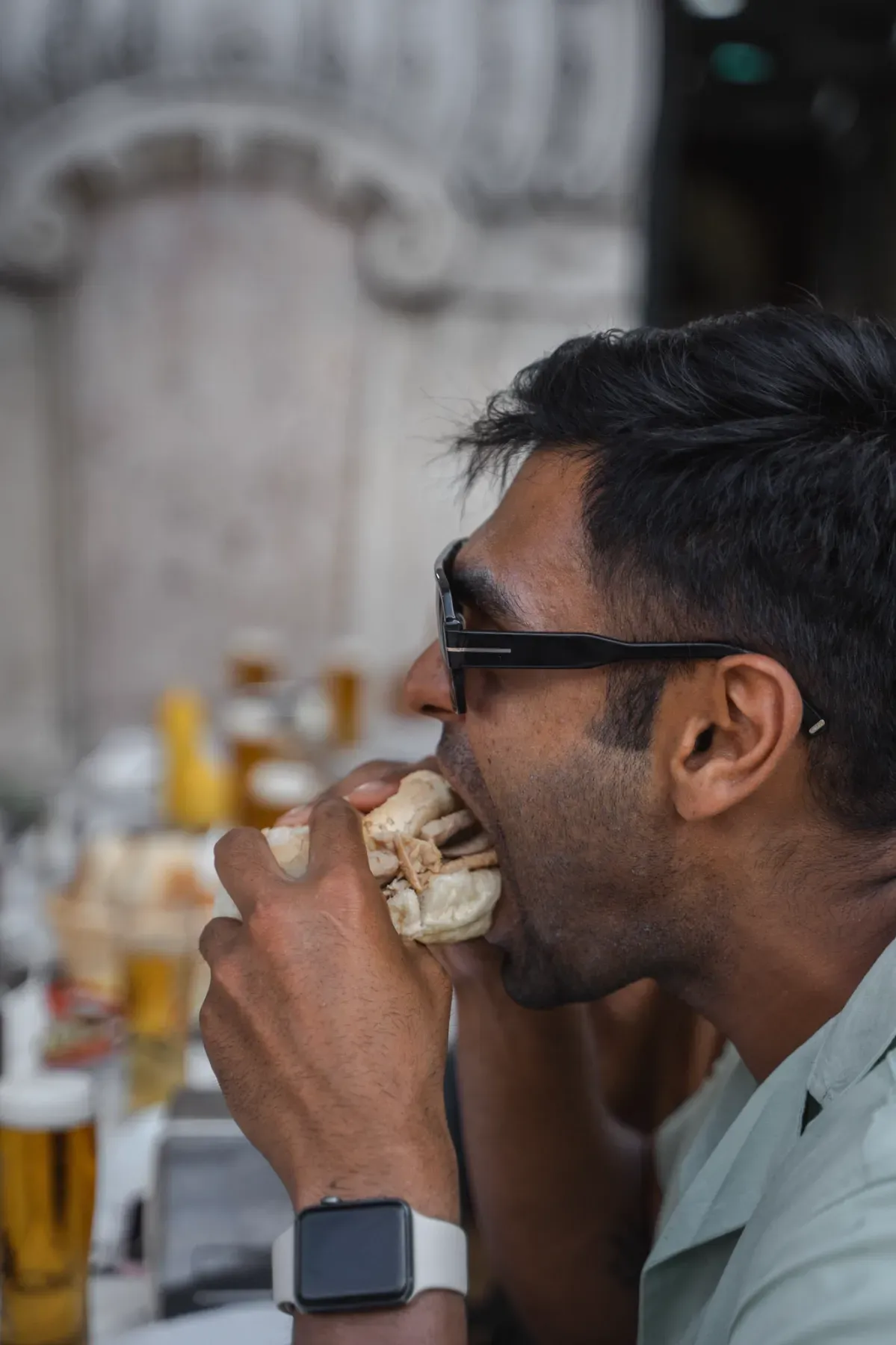 Man in sunglasses enjoying a traditional Portuguese bifana sandwich at an outdoor cafe in Lisbon, with a glass of beer on the table.