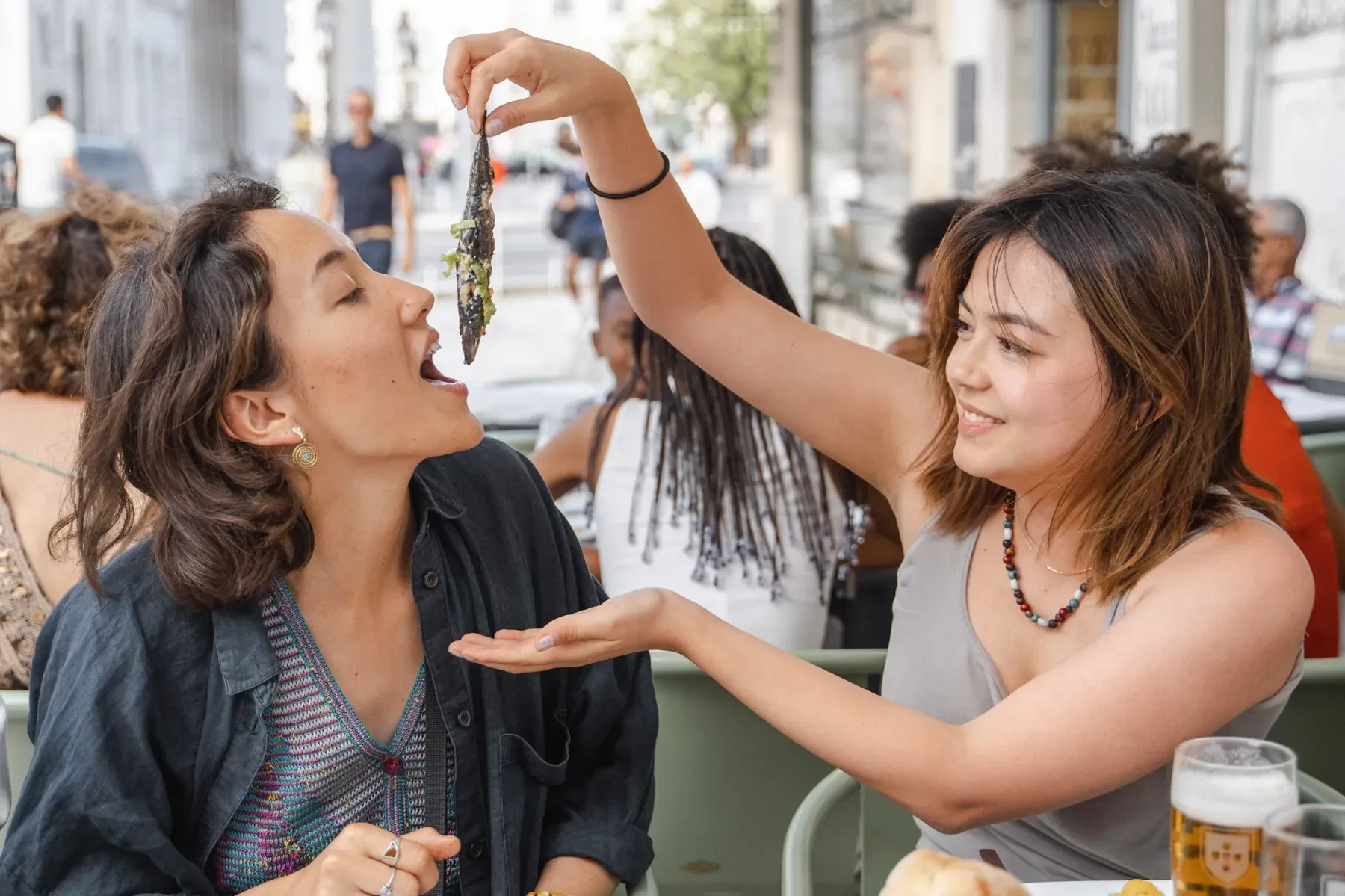 Two women at an outdoor cafe, one feeding the other a whole grilled sardine, a popular Portuguese delicacy.