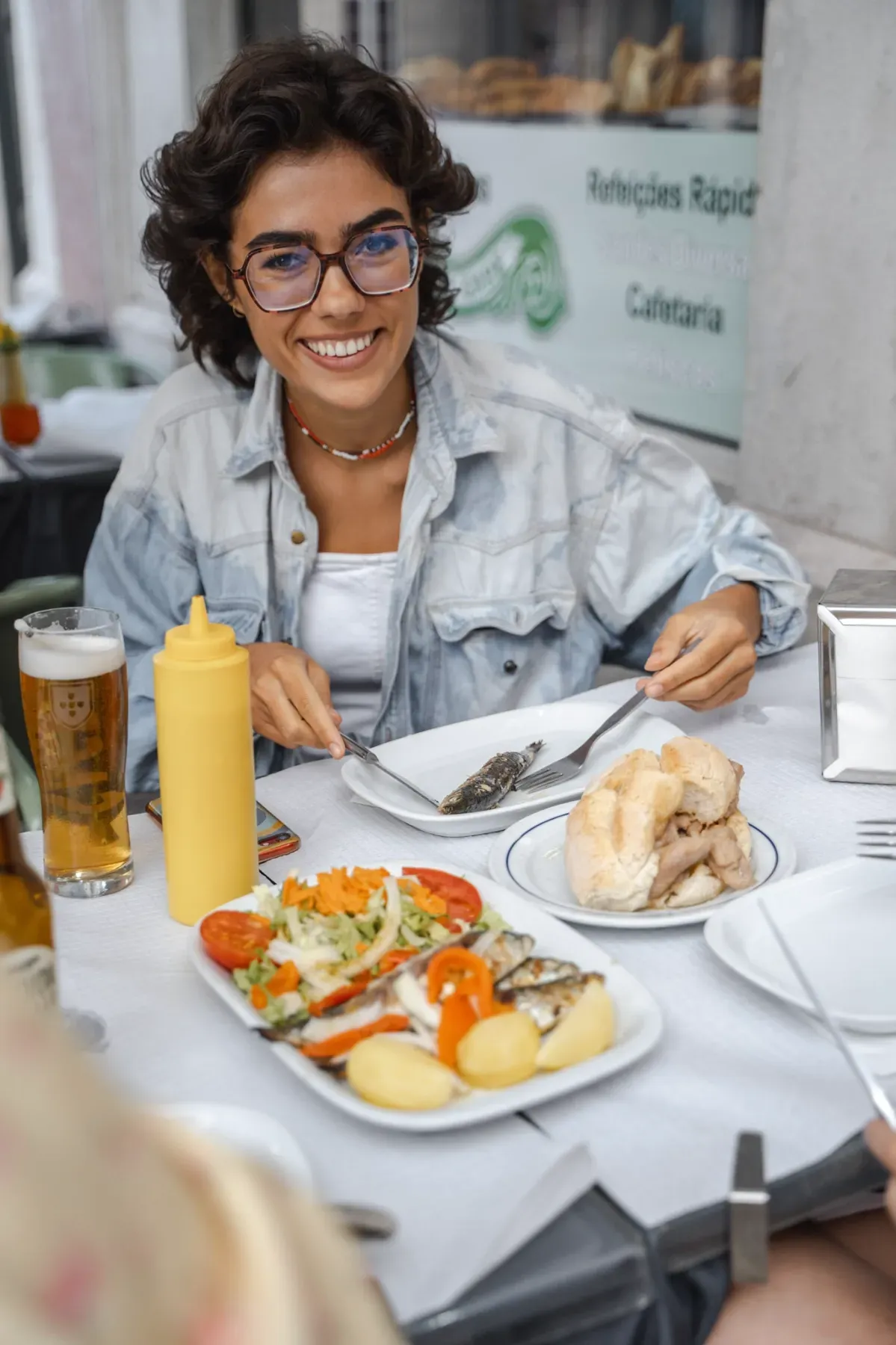 Happy woman with glasses eating grilled Portuguese sardines and salad with a beer at a Lisbon cafe.