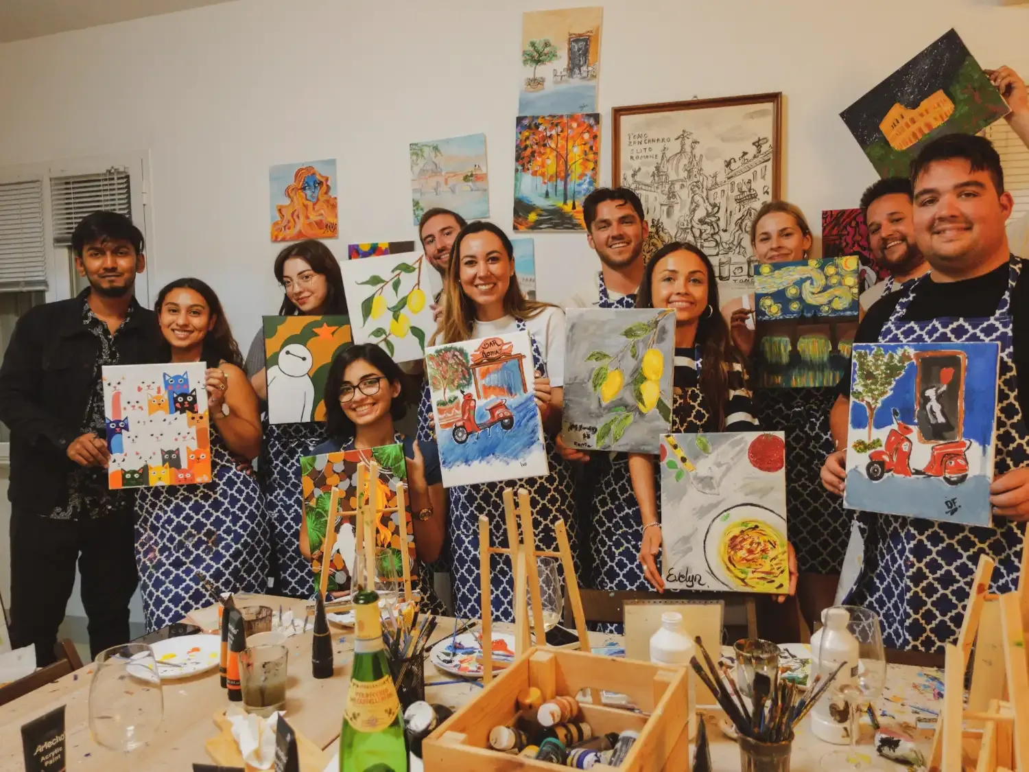 Large group photo of all participants smiling with their finished artwork displayed, wine bottles on table