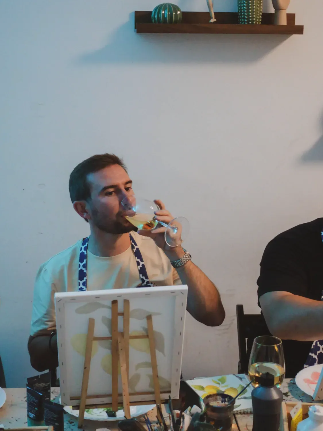 Man enjoying wine while painting on canvas with art supplies and snacks on table during tipsy art class