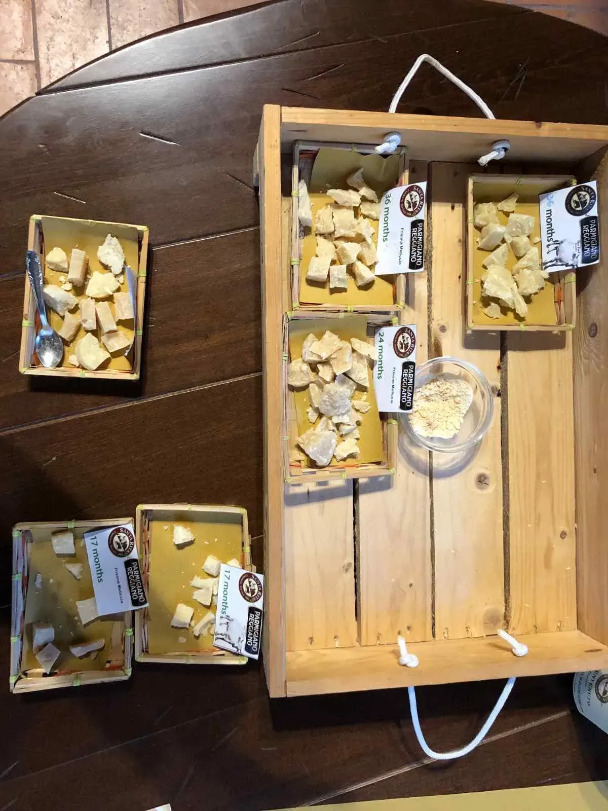 A wooden tasting display featuring multiple samples of aged Parmigiano Reggiano cheese in labeled boxes with varying ages and textures, arranged on a dark wooden table for a guided cheese tasting experience during the farm tour.
