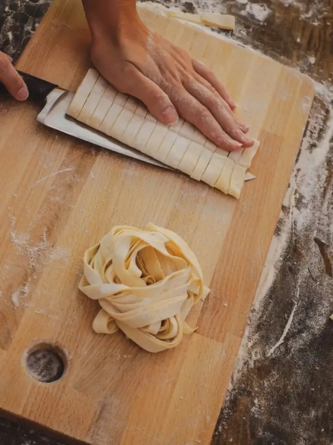 Close-up of hands cutting fresh homemade pasta dough into thin fettuccine strips on a wooden cutting board with flour