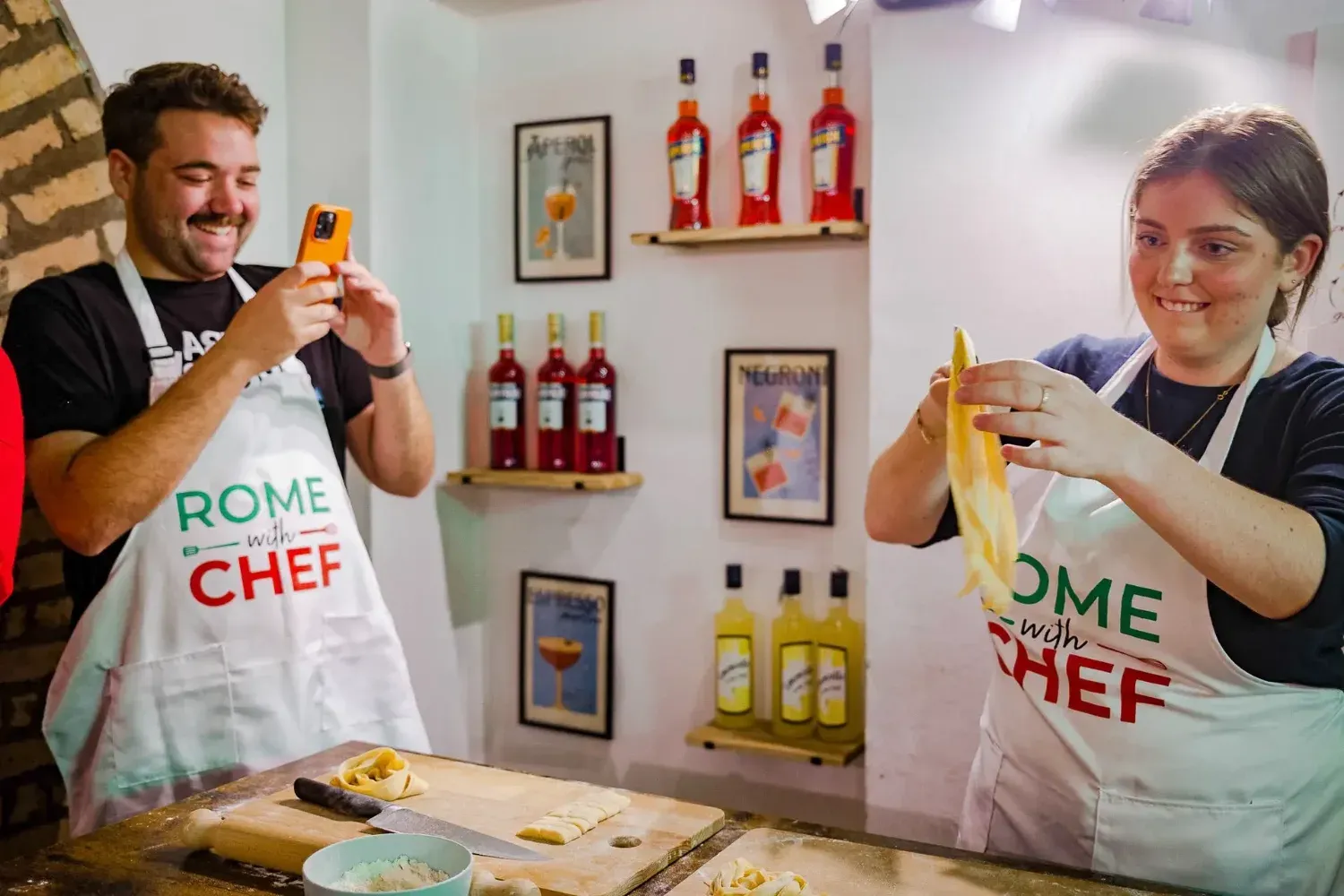 Man taking a photo of woman holding fresh pasta strands in Rome with Chef kitchen studio with wine bottles in background