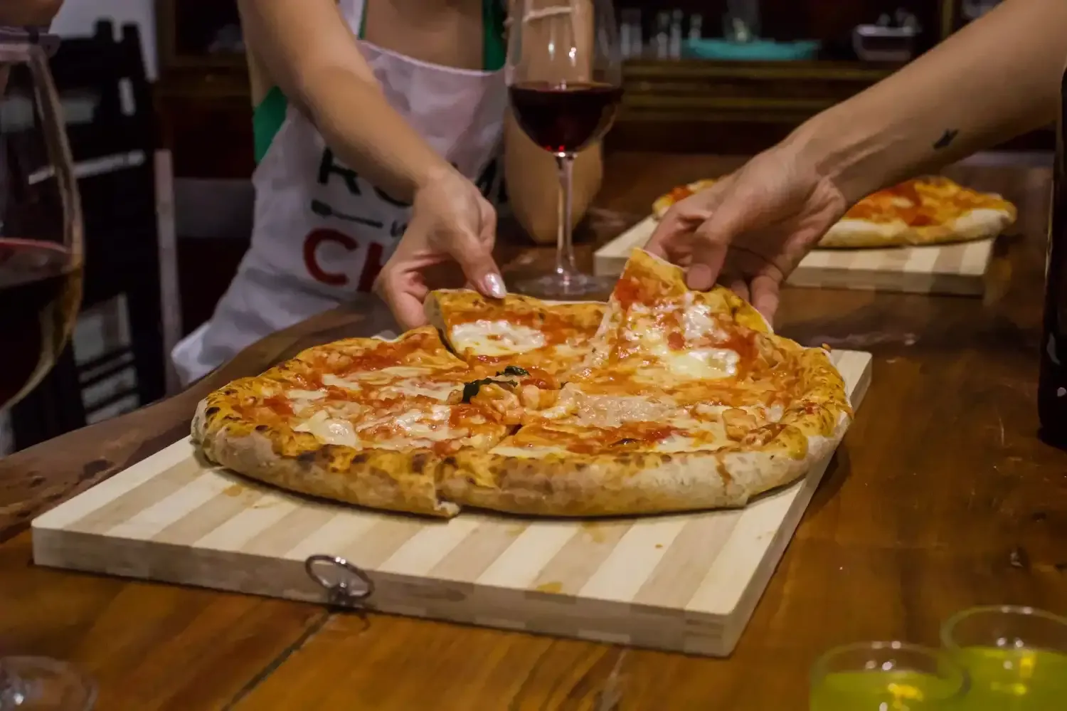 Close-up of hands breaking a freshly baked pizza with melted cheese and basil while holding wine glass