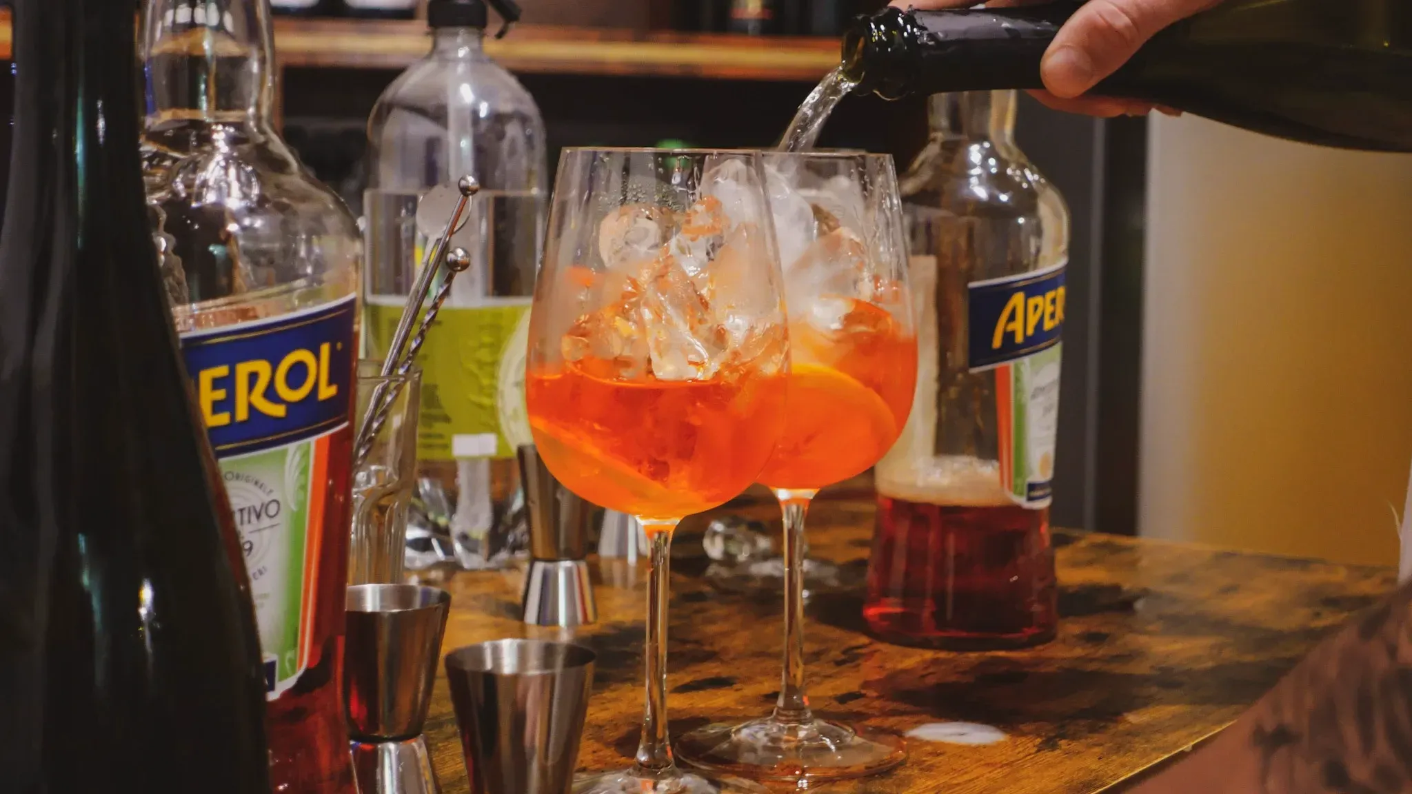 Close-up of cocktail bar setup with Aperol and Erol bottles, cocktail shaker, and vibrant red Spritz drinks being prepared