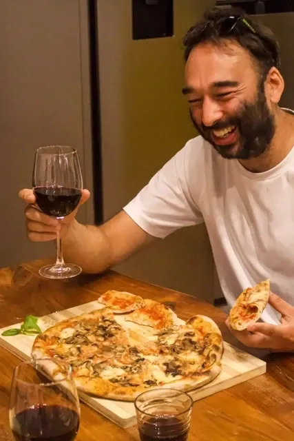 Happy man toasting with a glass of red wine while enjoying a freshly made pizza with melted cheese during the cooking class