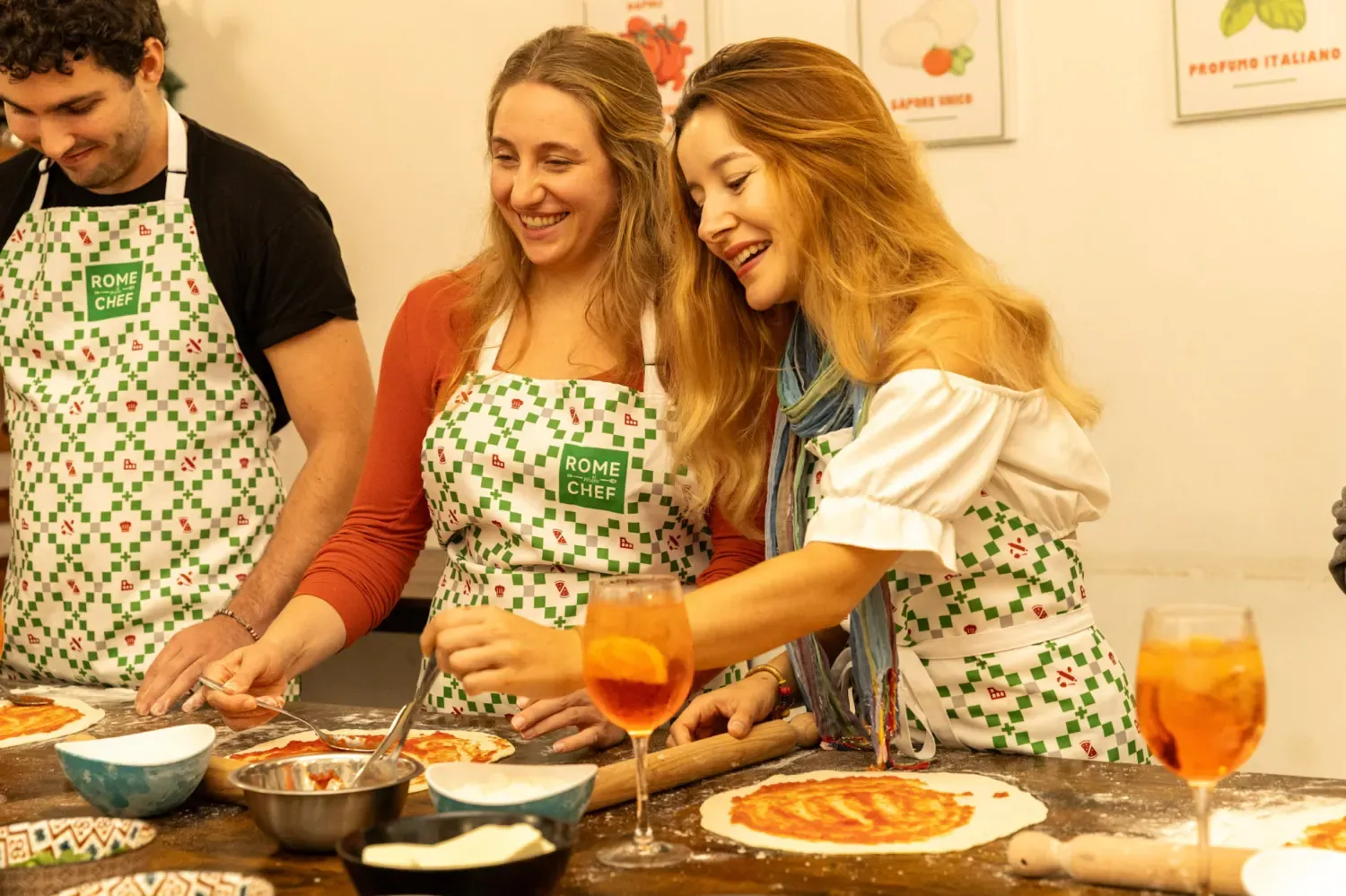 A group of happy participants wearing Rome with Chef aprons enjoying cocktails while learning to make authentic Italian pizza during a hands-on cooking class in Rome, with fresh pizza and ingredients on the counter.
