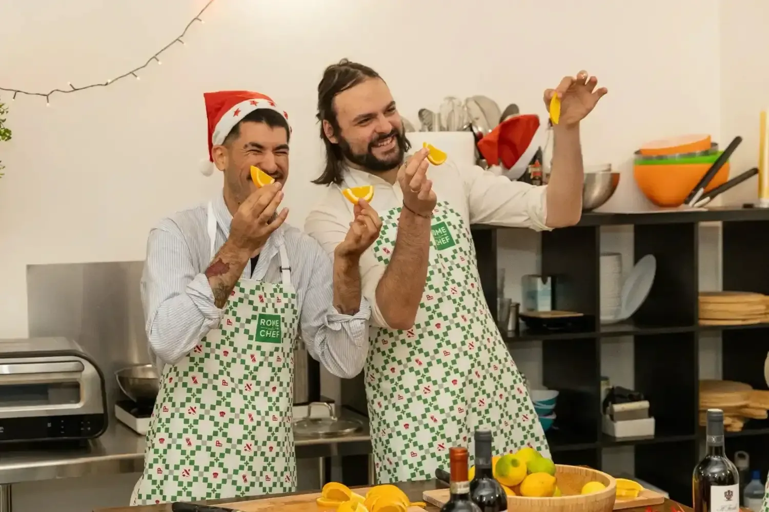 Two men in Rome with Chef aprons and Santa hats playfully holding orange slices in their mouths during cooking class