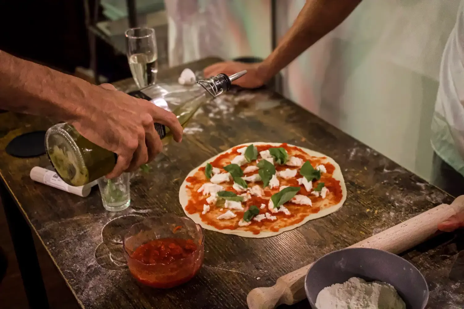 Hand pouring white wine into a glass next to a freshly made Margherita pizza with basil and mozzarella