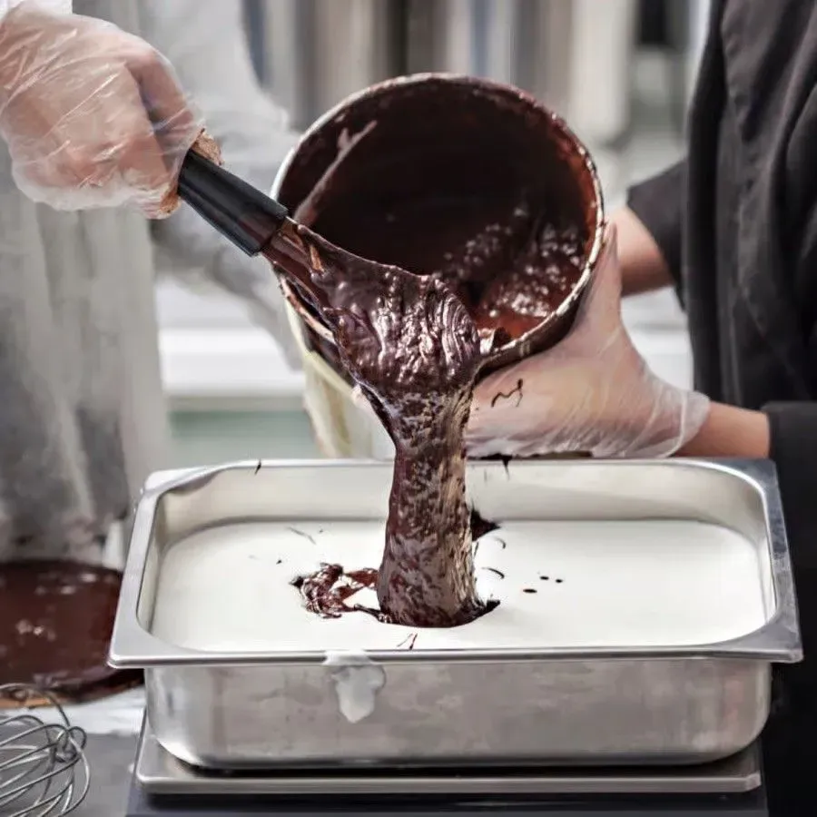 Hands pouring rich, dark chocolate gelato mixture from a container into a metal pan during a hands-on gelato-making class, demonstrating the mixing and preparation technique.