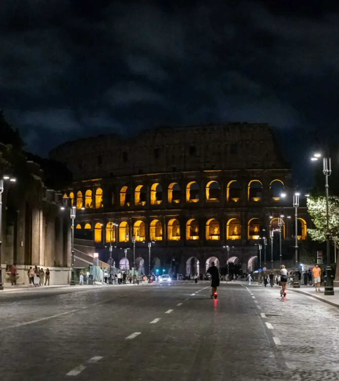 The ancient Colosseum amphitheater beautifully illuminated at night with tourists walking in the plaza