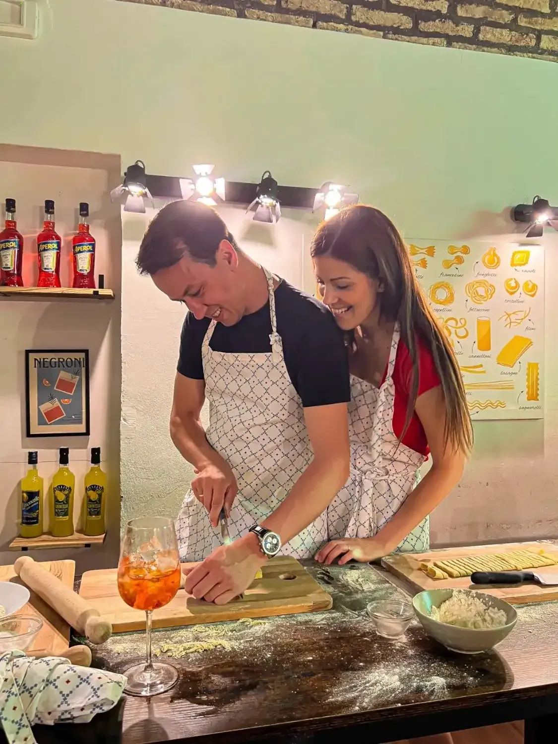 Couple in Rome with Chef aprons smiling while making fresh pasta with a Spritz cocktail on the table beside them