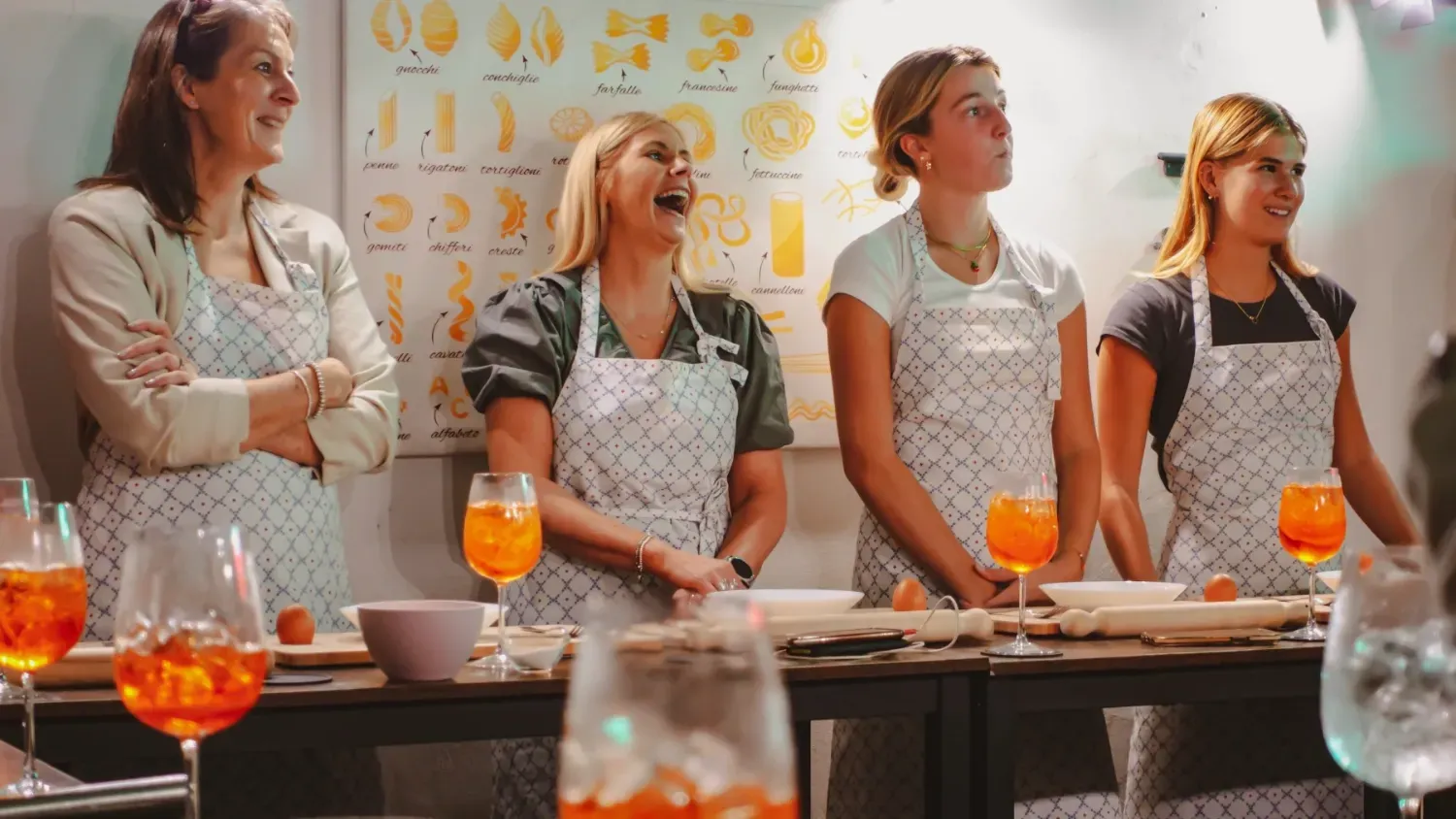 Group of four smiling women in Rome with Chef aprons holding Spritz cocktails in front of a pasta types chart