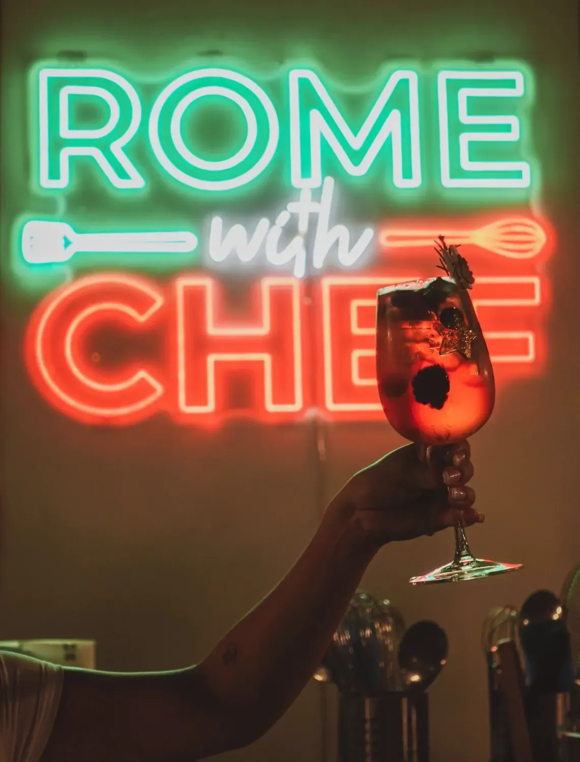 Hand holding a vibrant red Spritz cocktail in front of a neon Rome with Chef sign in the kitchen studio