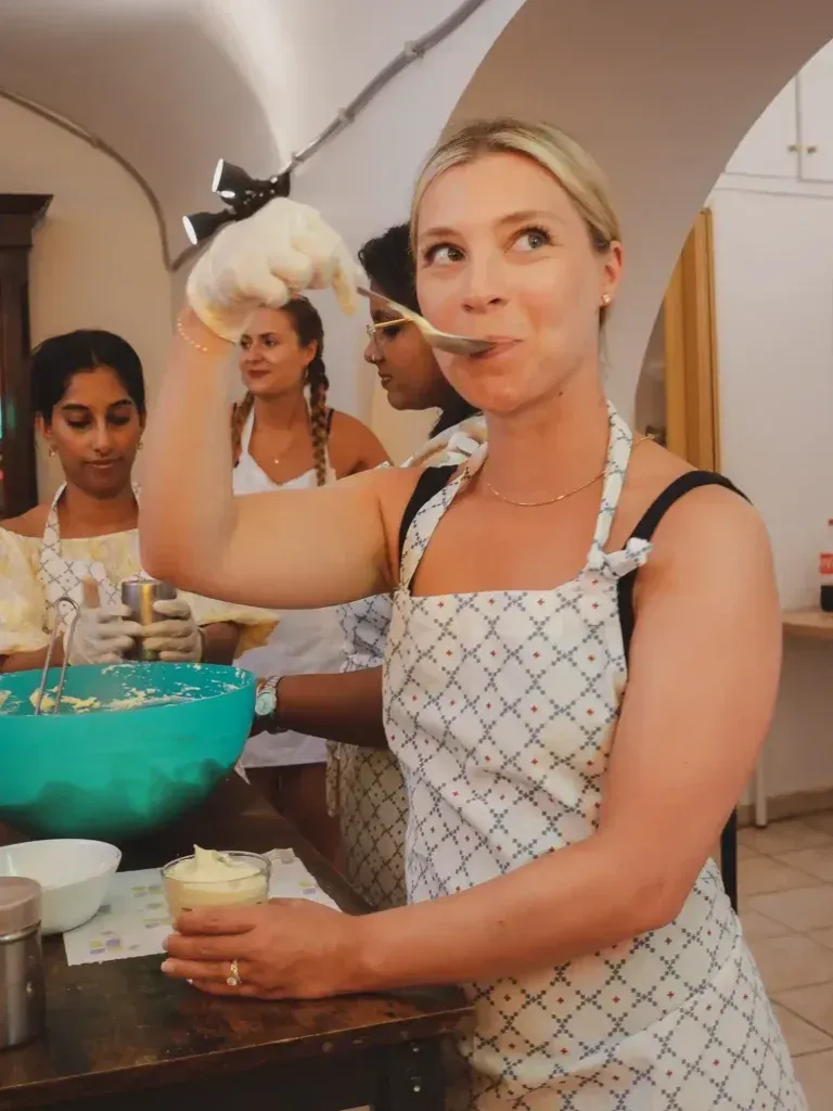 Woman in an apron preparing tiramisu, holding a whisk and a bowl in a kitchen setting.