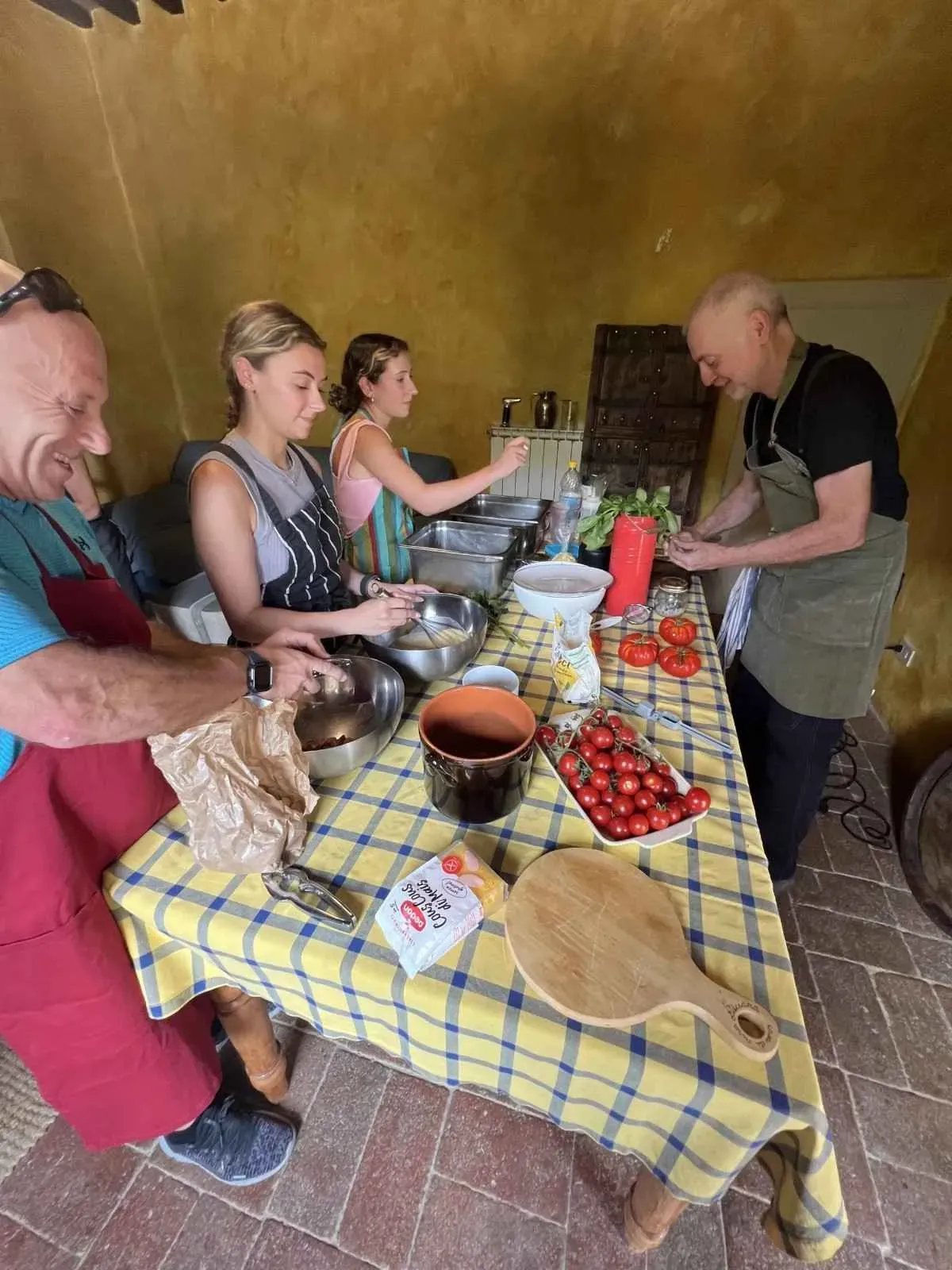 A chef instructing a group of tourists in a rustic yellow kitchen during a hands-on Tuscan cooking class, with fresh ingredients and cooking equipment on the counter.