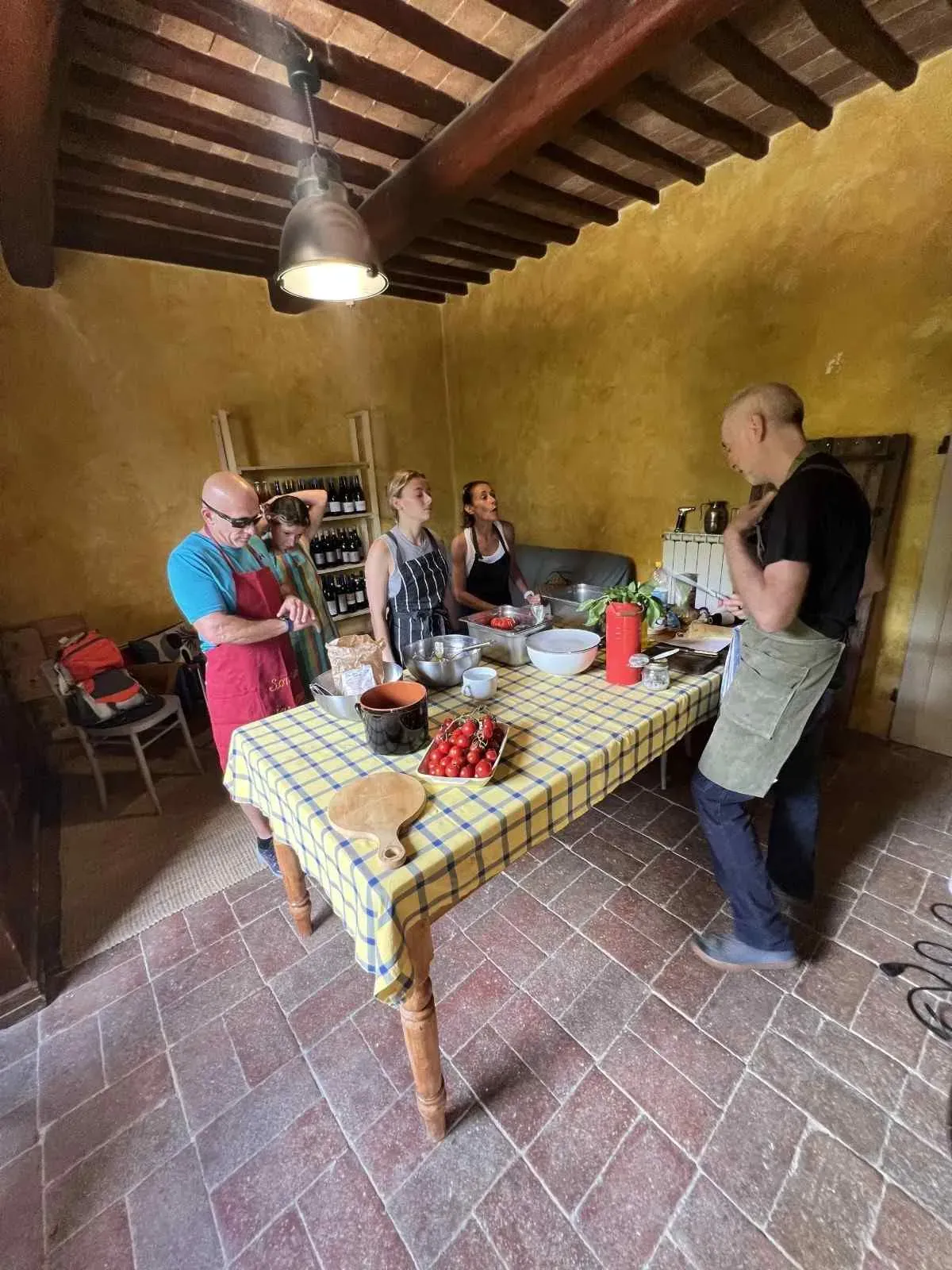 A chef instructing a group of tourists in a rustic yellow kitchen during a hands-on Tuscan cooking class, with fresh ingredients and cooking equipment on the counter.